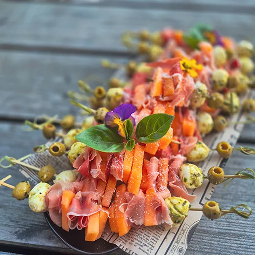 Vorspeisen-Spieße mit Melone, Parmaschinken, Mozzarella und Oliven auf Zeitungspapier – Antipasti von Kostbarkeiten